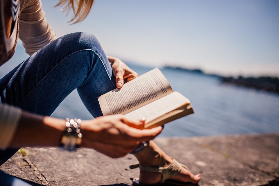 foto waarop een been te zien is en een hand met een open boek - met op de achtergrond zand en water
