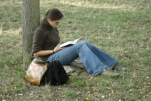 Vrouw zit te lezen in het gras met haar rug tegen een boomstam