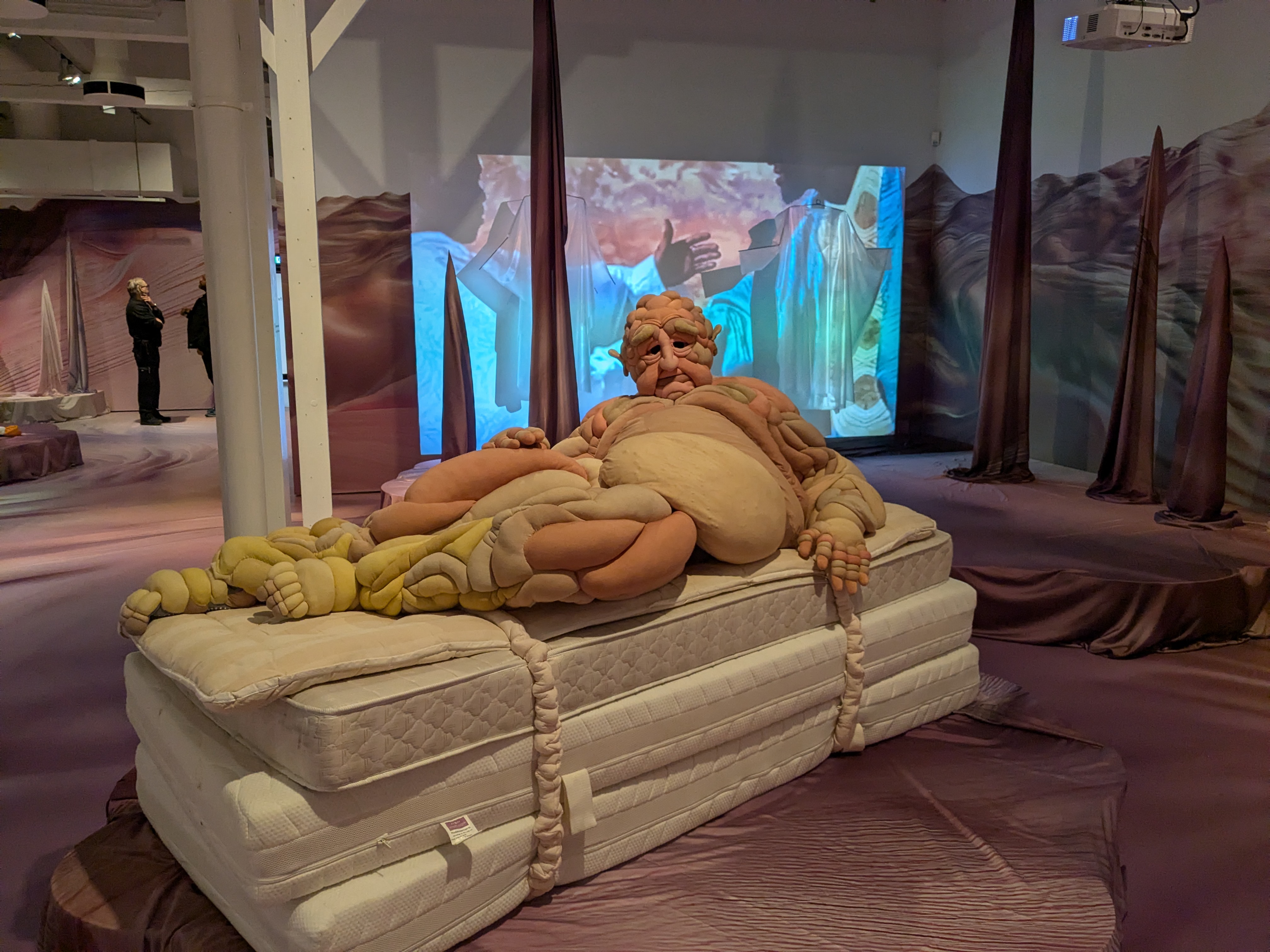 De afbeelding toont de kunstinstallatie ‘Dave’ in een modetentoonstelling van het Textielmuseum. Op de voorgrond ligt een groot, sculpturaal mannelijk figuur op een stapel matrassen. Dit figuur heeft een uitvergroot, vervormd lichaam met veel huidplooien, gemaakt van textiel in zalmroze, gele en beige tinten. Het gezicht van de figuur heeft diepe rimpels en hangende oogleden. De handen rusten op het lichaam en bestaan uit hetzelfde zachte, opgevulde materiaal. 