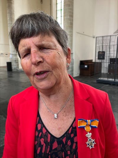 Portret van een trots kijkende Ineke Glorie in een rode blazer, met op haar linker-rever haar koninklijke onderscheiding