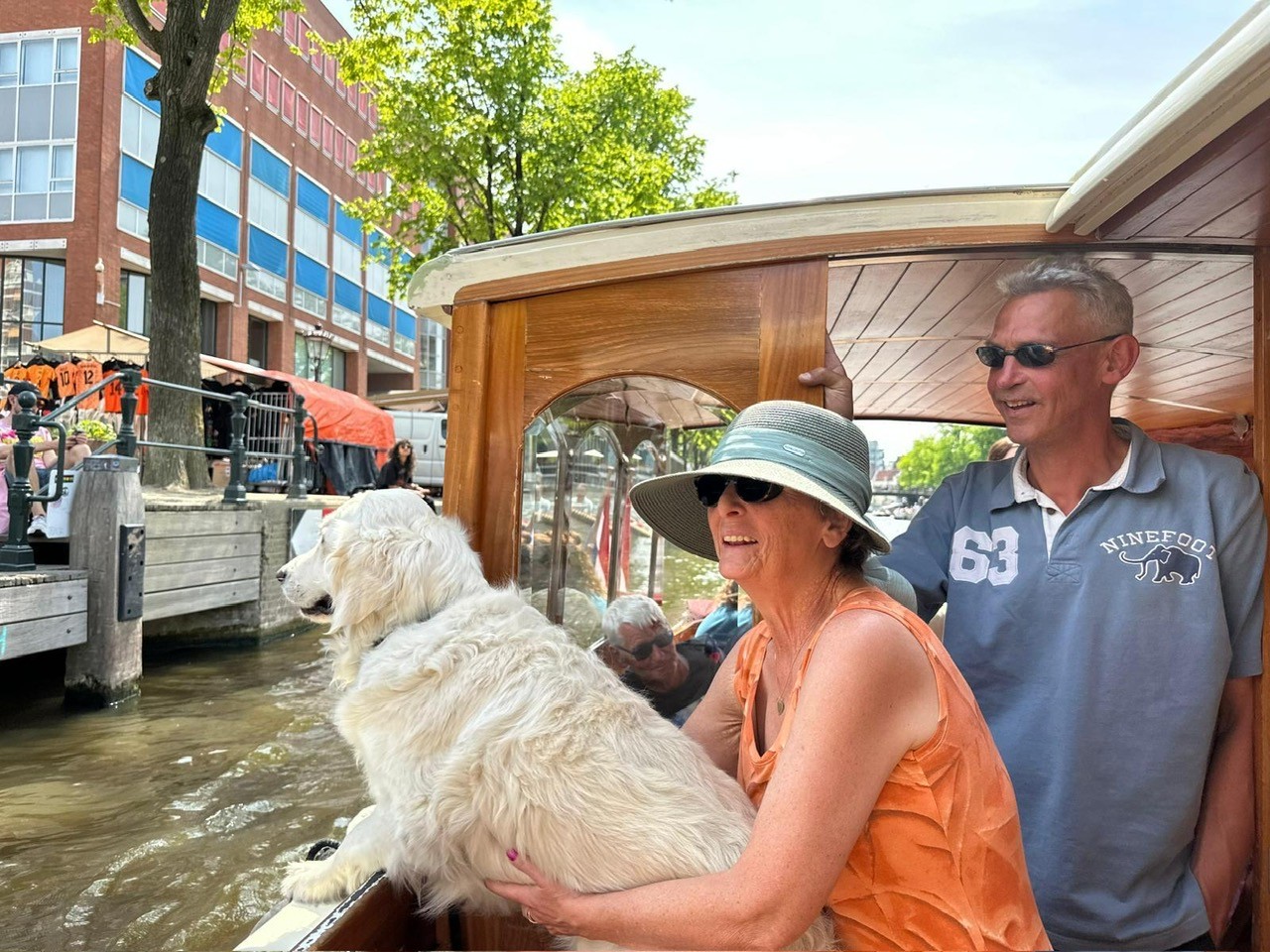 Sabrina Heemskerk die op een bootje langs een volle kade vaart. Ze leunt op een golden retriever, die op zijn of haar beurt op de rand van de boot leunt. Achter Sabrina staat een man. Hij draagt een zonnebril. Het is een mooie dag. Sabrina draagt een oranje mouwloos shirt en een visserhoedje. 