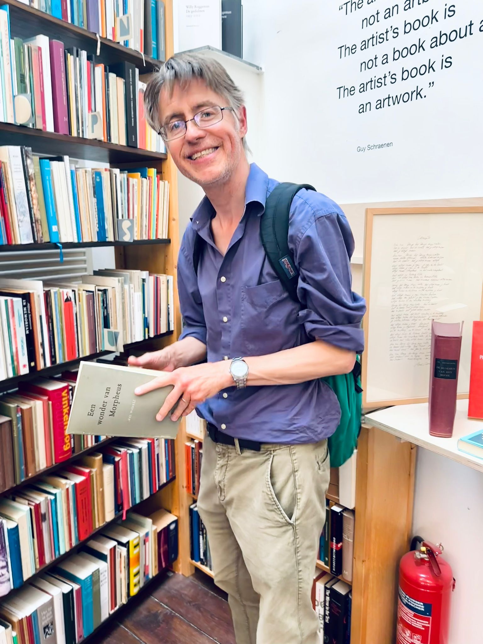 Guido Spring met een rugzak op zijn rug en een brede glimlach om zijn mond. Hij bevindt zich tussen de boekenkasten. 