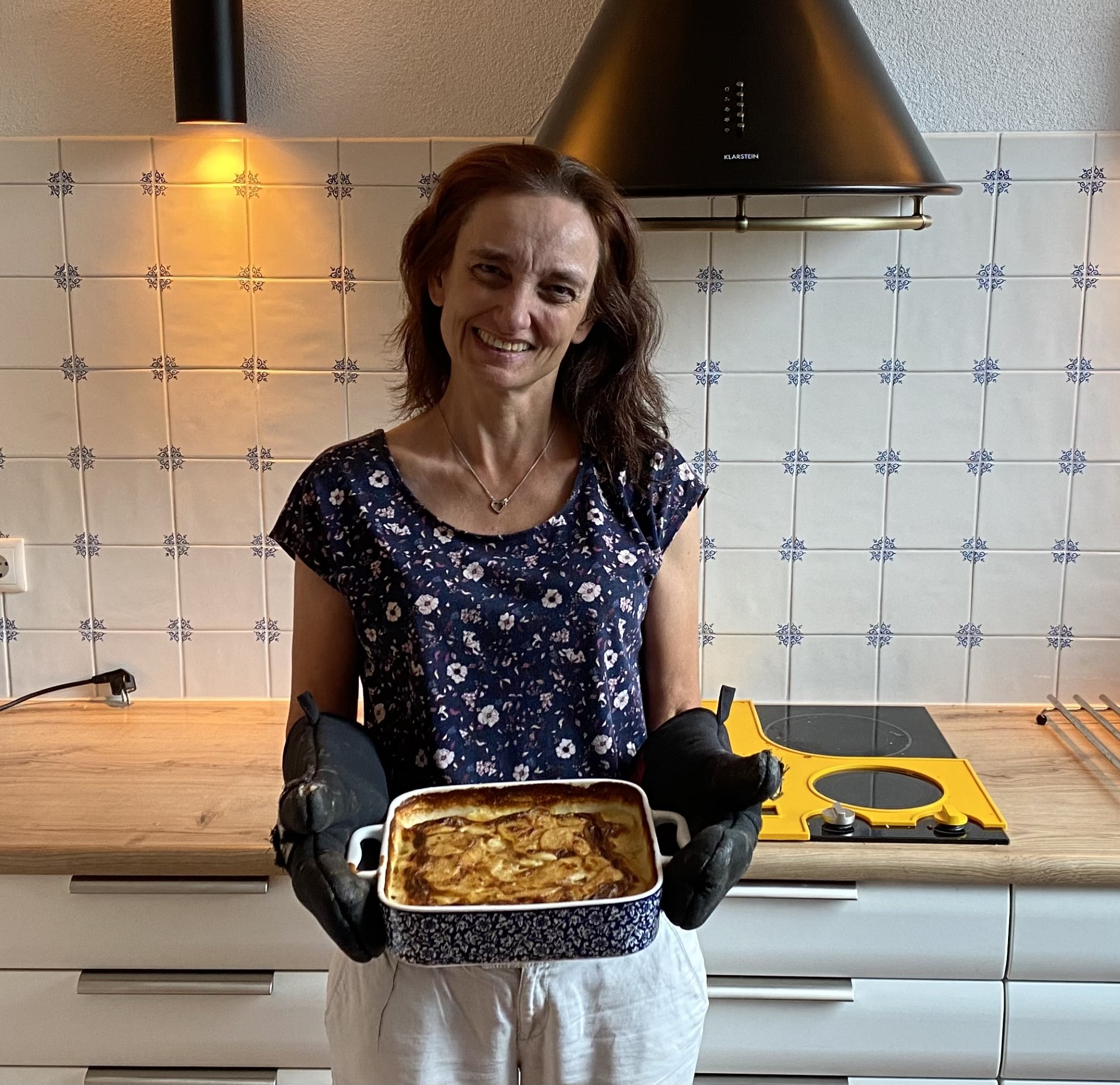 Marloes Schaap die lachend de camera inkijkt. Ze bevindt zich in een keuken en in haar handen heeft ze een ovenschaal. De inhoud ervan toont ze aan de kijker.