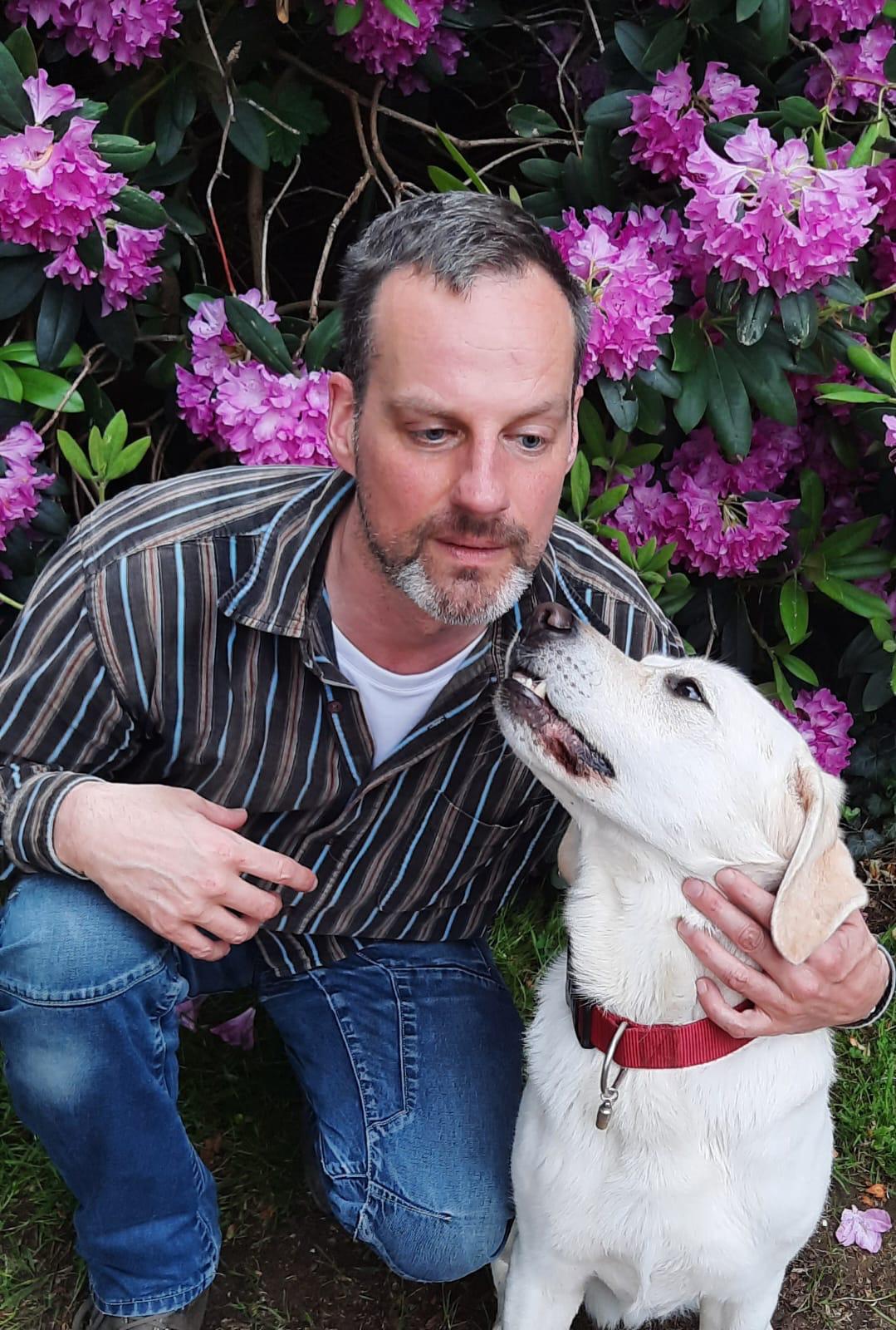 Marcel van der Bie, knielend voor een struik met roze bloemen met links van hem een blonde labrador die naar hem opkijkt