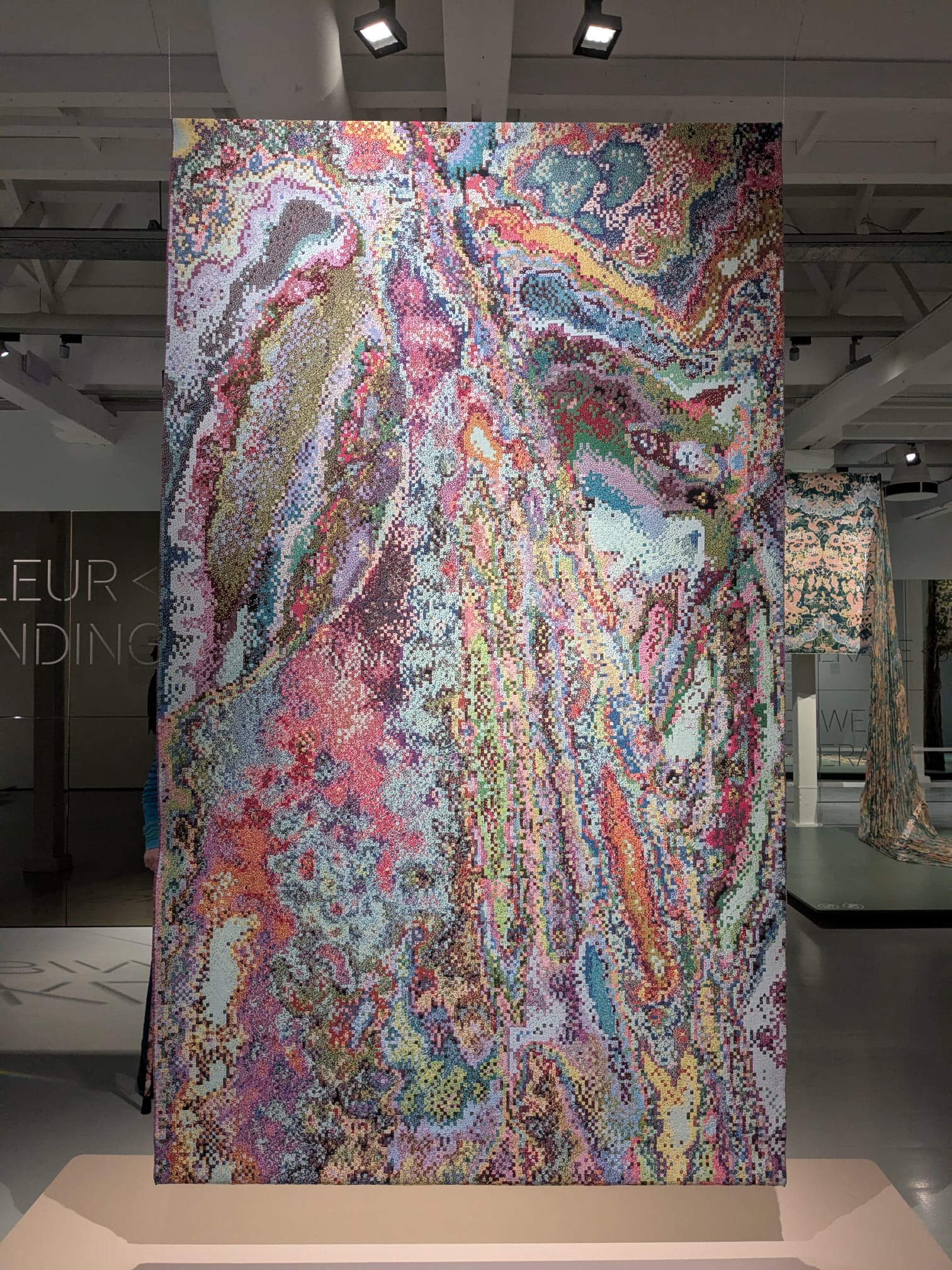 Een kunstwerk uit de expositie in het TextielMuseum. Het is een groot kleed van textiel waarop allerlei kleuren door elkaar gemengd zijn.