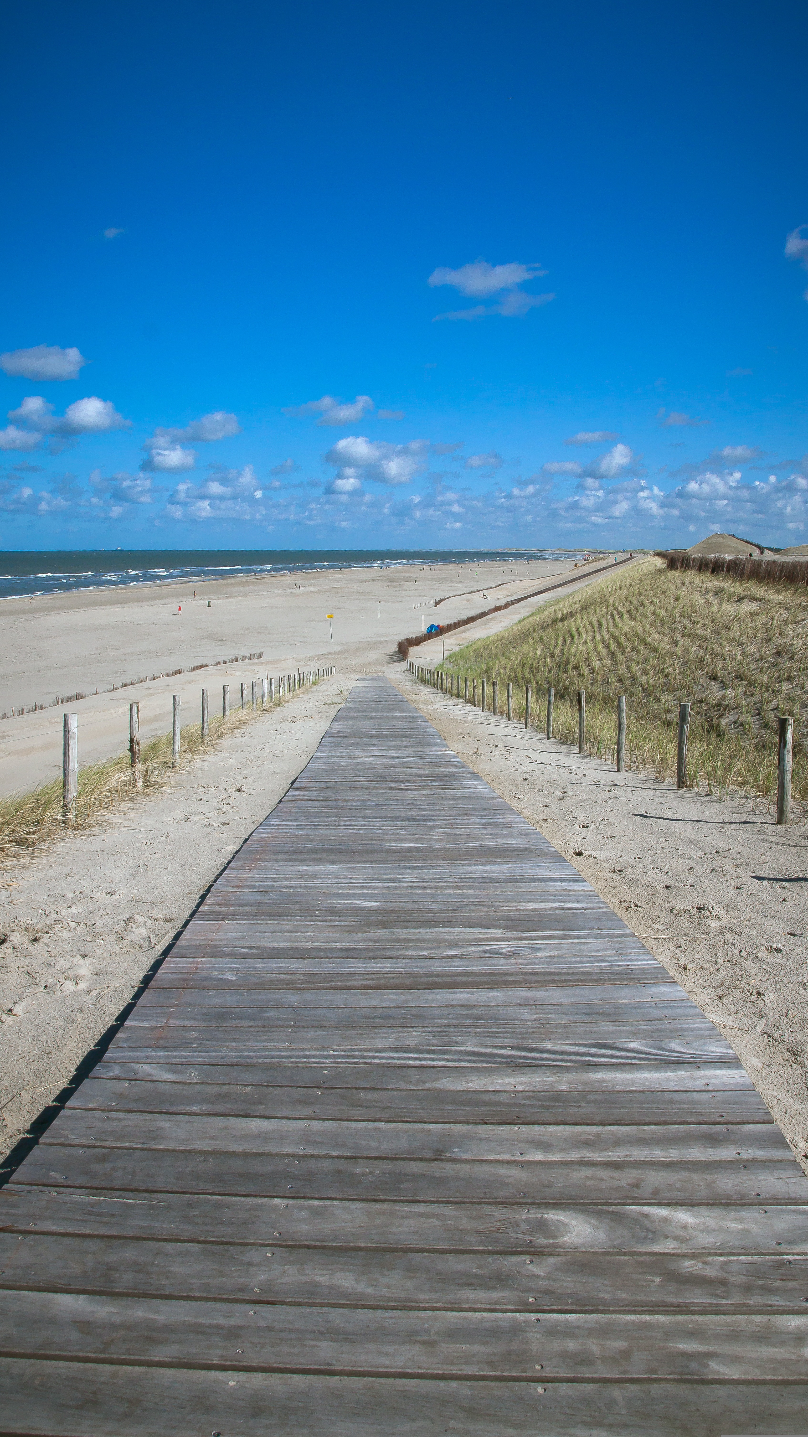 Een strand gezien vanaf een vlonderpad. Het pad lijkt steeds smaller te worden naarmate het verdwijnt in de achtergrond. Aan de rechterkant van het vlonderpad zijn duinen te zien, aan de linkerkant een leeg strand met in de verte de zee. De bovenkant van de foto wordt gedomineerd door een strakblauwe lucht met her en der wat verdwaalde witte wolken. 