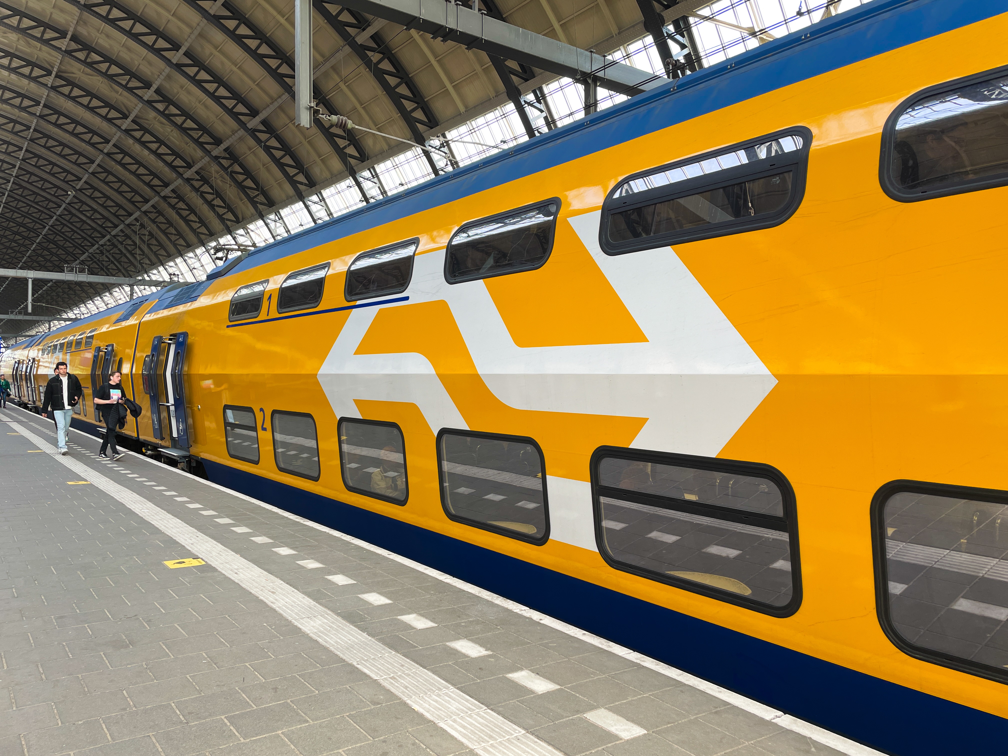 Deel van een overdekt perron waarlangs een stilstaande trein. Het perron is verlaten op twee mensen na die richting de openstaande treindeuren lopen.