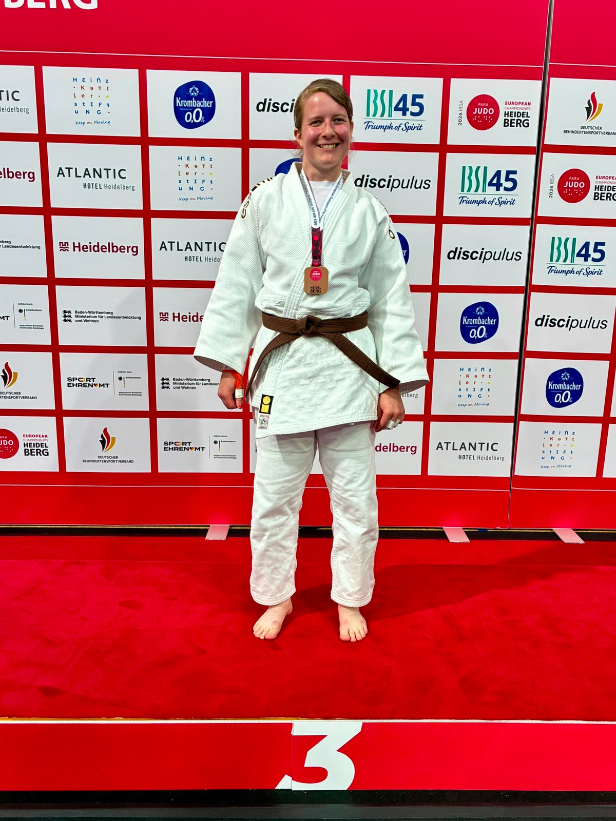 Larissa Klaassen in wit judopak met bruine band. Ze staat voor een muur met afbeeldingen van verschillende sponsors en draagt een bronskleurige medaille om haar nek. Ze staat op een rode ondergrond met onderaan het cijfer 3. 