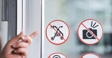 Hand die wijst naar een sticker op een raam. Op de sticker staat in een pictogram aangegeven: verboden voor rollators