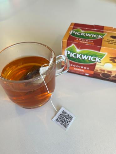 Glazen kopje waar een theezakje in hangt. Het labeltje aan het zakje rust op een tafelblad en laat een QR code zien. Naast het kopje staat een doosje rooibosthee