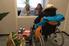 Inkijkje in de portacabin, op de voorgrond een dame in een rolstoel, met naast haar bloemen en lekkernijen. aaan de andere kant van het glas een man en vrouw, pratend in een microfoon.