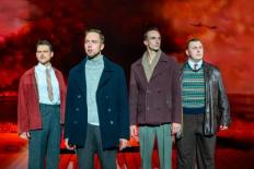 Scenefoto van vier ernstig kijkende mannen op het toneel. Ze staan naast elkaar tegen een rodegekleurde achtergrond van rook en propellorvliegtuigen.